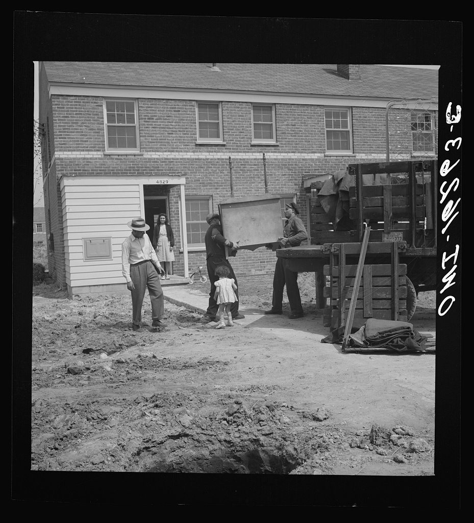 Detroit, 1942. A Black family moves into the Sojourner Truth homes. Two men lower a piece of furniture from the back of a truck; a small child in a white dress stands by a tricycle on the paved walk, and a woman waits in the doorway of the unit. A man in a fedora stands on the churned ground in the foreground. The brick housing block is behind them.