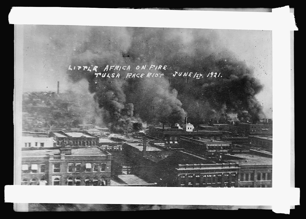 Black-and-white photograph of the Greenwood district of Tulsa, Oklahoma, burning on 1 June 1921. Heavy smoke rises from blocks of two- and three-storey brick buildings. A caption has been written by hand directly into the negative, in two lines: "Little Africa on fire / Tulsa race riot June 1st. 1921."