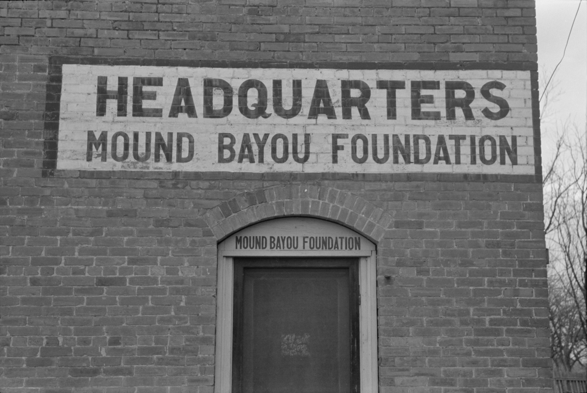 A brick building photographed straight on. A painted sign across the upper wall reads HEADQUARTERS in large letters with MOUND BAYOU FOUNDATION beneath it. A smaller board above a panelled doorway repeats MOUND BAYOU FOUNDATION. Bare winter branches and a utility line at right.