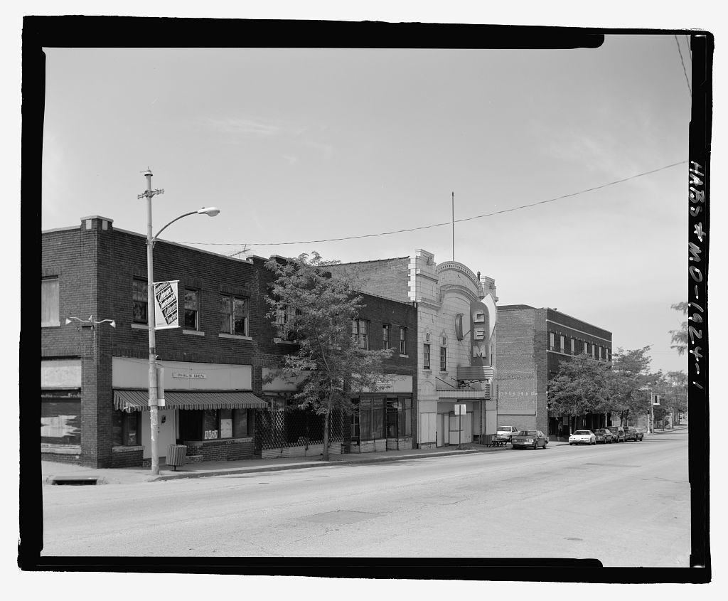 Black-and-white photograph of East Eighteenth Street, Kansas City, Missouri. A row of two-storey brick storefronts runs along the sidewalk under a street lamp; the vertical marquee of the Gem theater stands out against the sky. Cars are parked along the kerb. The street is intact and in use.
