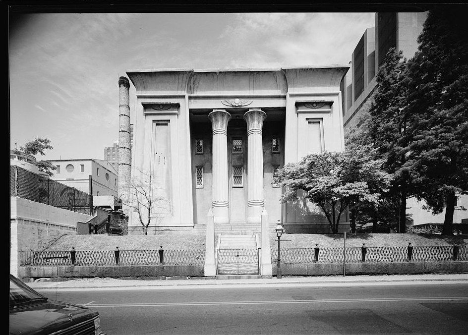 Black-and-white photograph of the Egyptian Building in Richmond, Virginia, seen straight on from across the road. An Egyptian Revival facade with two lotus-capital columns set between heavy battered piers, a cavetto cornice above, and a low iron fence along the sidewalk. A parked car sits at the lower left of the frame.