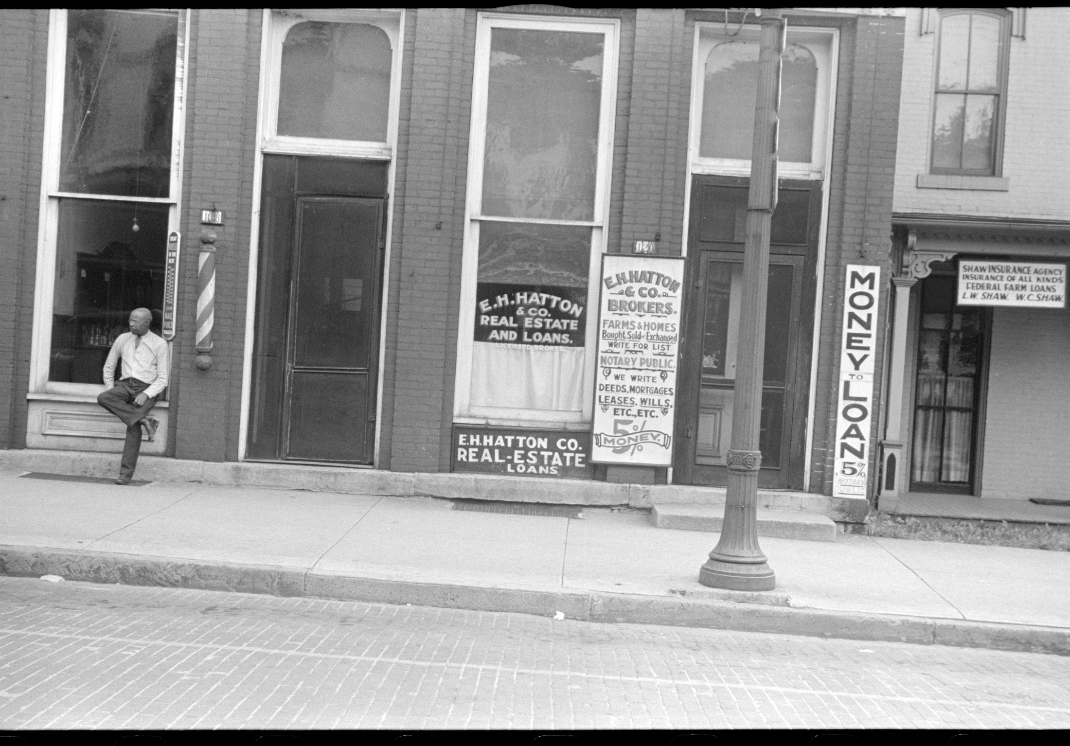 A brick storefront in Marysville, Ohio, summer 1938. Painted signs across the windows read E. H. Hatton & Co., Real Estate and Loans; Money to Loan, 5 percent; We Write Deeds, Mortgages, Leases, Wills; Farms and Homes Bought, Sold or Exchanged. A Black man in glasses, shirt and tie leans against the sill of the adjoining storefront, beside a barber pole.
