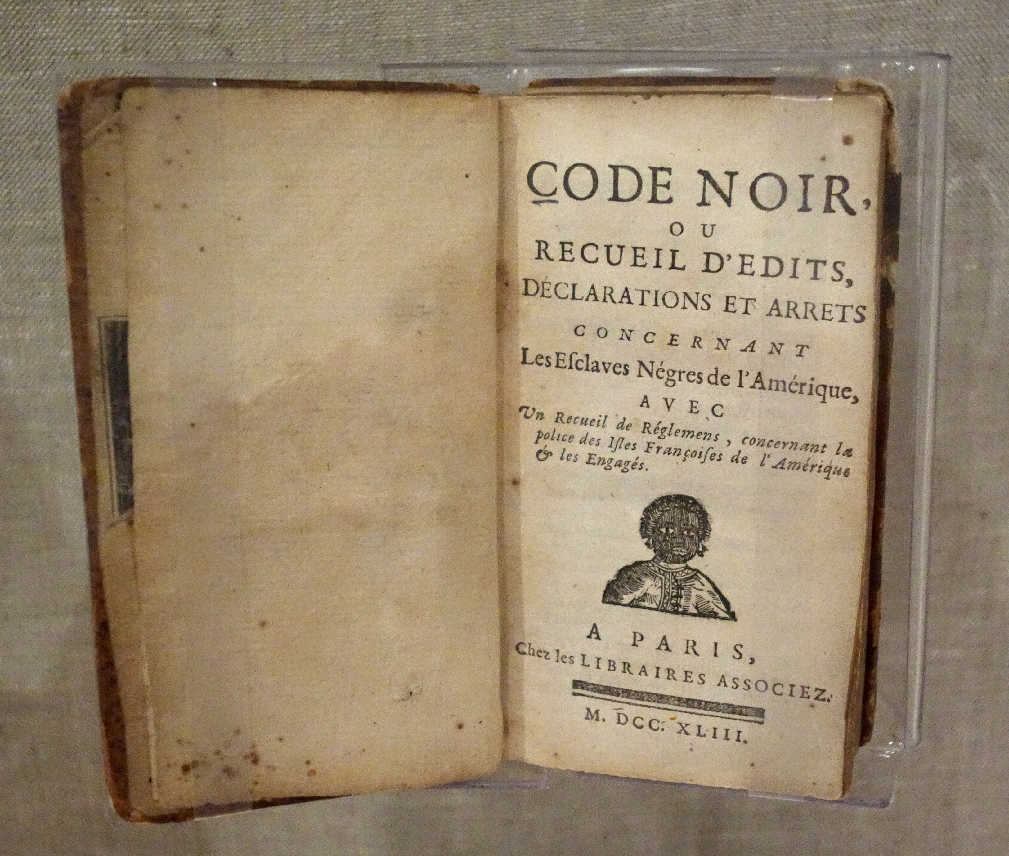 A small leather-bound volume lies open. The title page reads CODE NOIR, ou RECUEIL D'EDITS, DECLARATIONS ET ARRETS CONCERNANT Les Esclaves Négres de l'Amérique. Beneath it: A PARIS, Chez les Libraires Associez. M.DCC.XLIII.